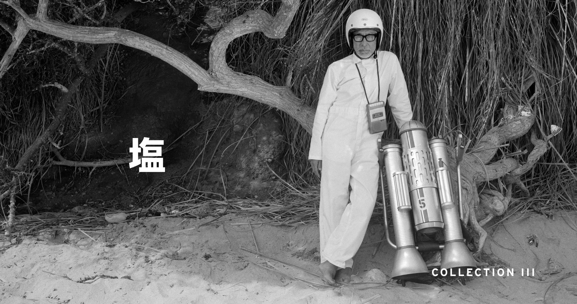 Black and white vintage photograph showing a person in a light-colored outfit and helmet standing outdoors near a jet pack wearing class eyewear